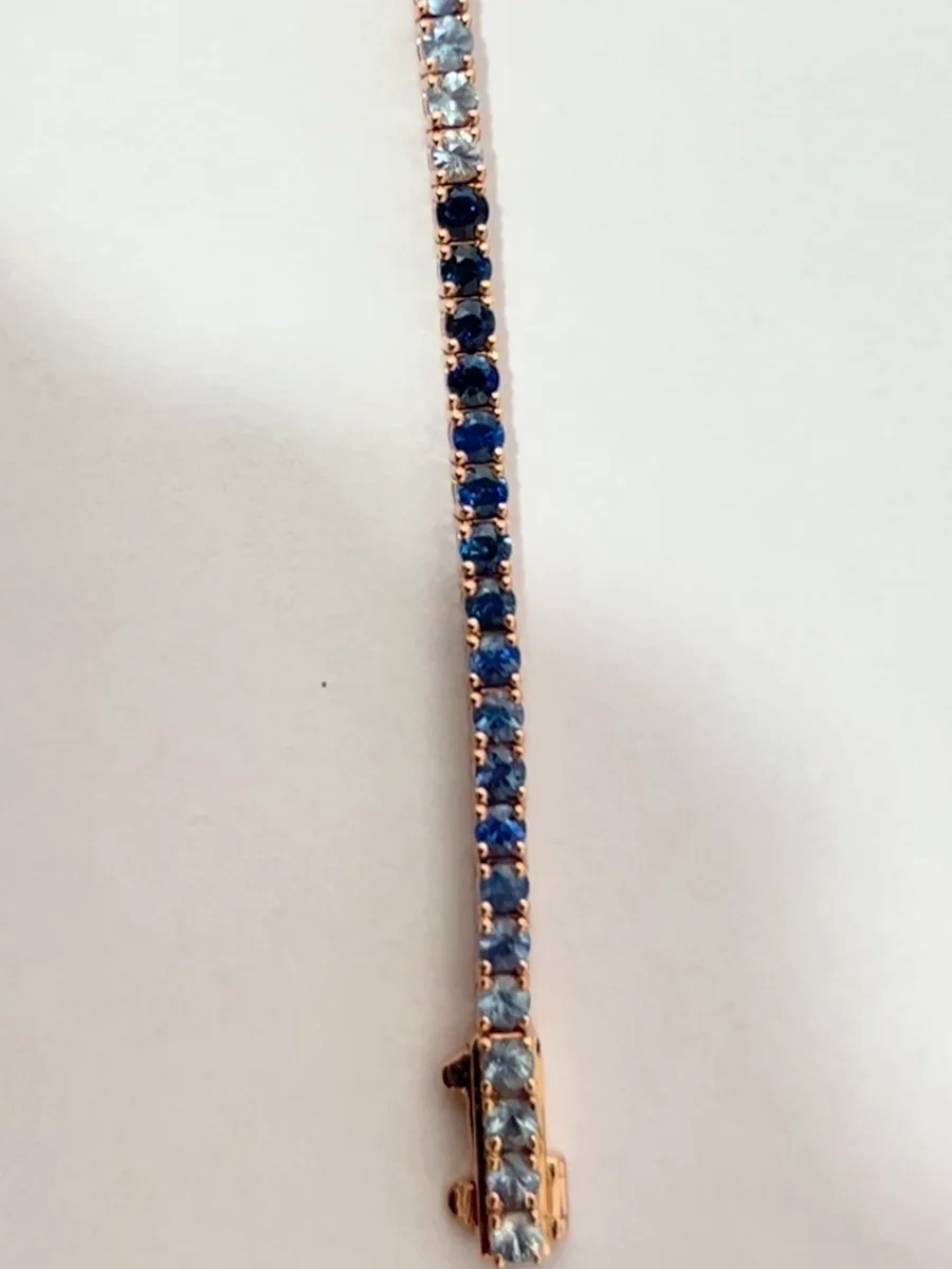 Close up of a 14K rose gold tennis bracelet: pale lab grown sapphires, a reset to deep blue, then a fade to pale stones set across the box clasp