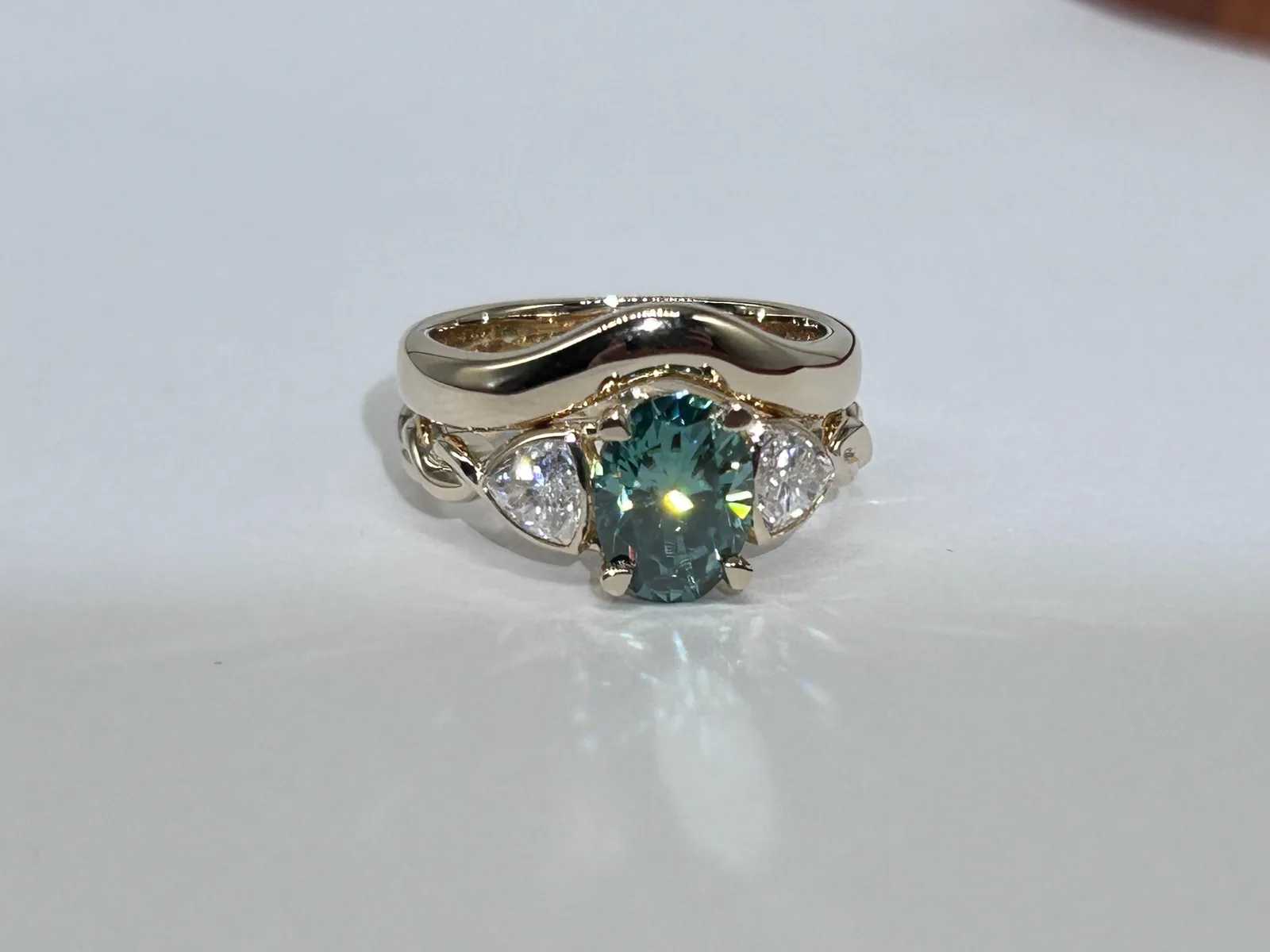 14K yellow gold three stone engagement ring with a teal oval center and two trillion side stones, worn with a polished wedding band that curves over the center stone